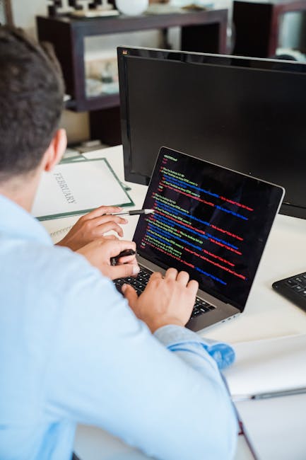A person sitting at a desk working on a laptop computer displays a screen filled with lines of colorful code, with a large external monitor positioned directly in front of them. The individual is wear