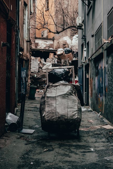 Three large black refuse bags made of thick, glossy plastic material are positioned on a section of paved pavement adjacent to a curb during daytime. The bags appear tightly filled and are leaning aga