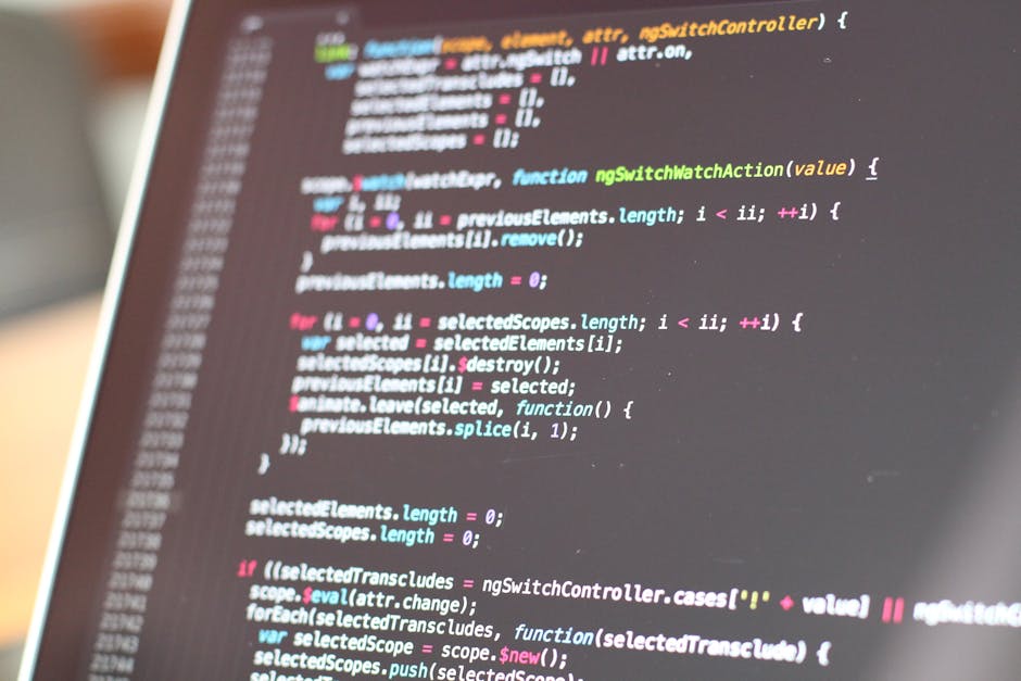 A close-up of a person's hands typing on a laptop keyboard, which displays a dark-themed coding interface with colorful lines of code and text. The laptop is situated on a smooth, dark surface, with t